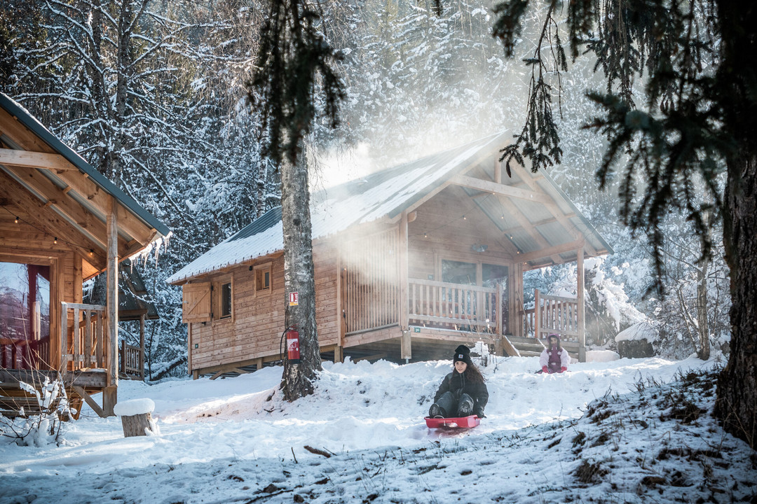 Deux enfants qui font de la luge devant leur chalet loué chez Huttopia pendant leurs vacances d'hiver à la montagne.