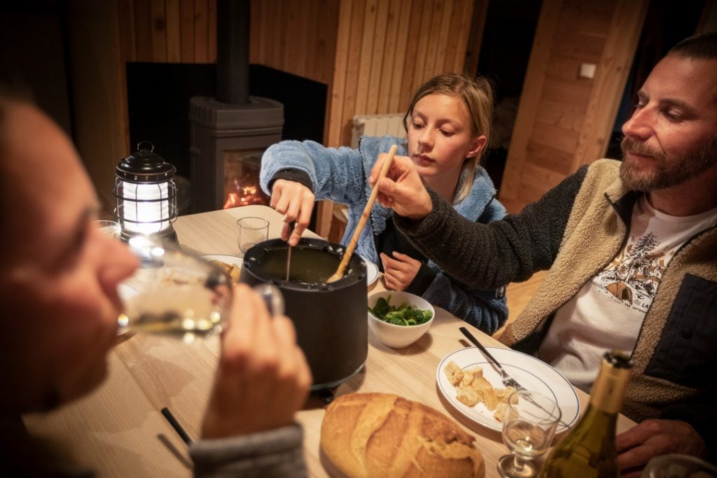 Famille qui mange une fondue dans leur chalet loué chez Huttopia pendant leurs vacances d'hiver à la montagne.